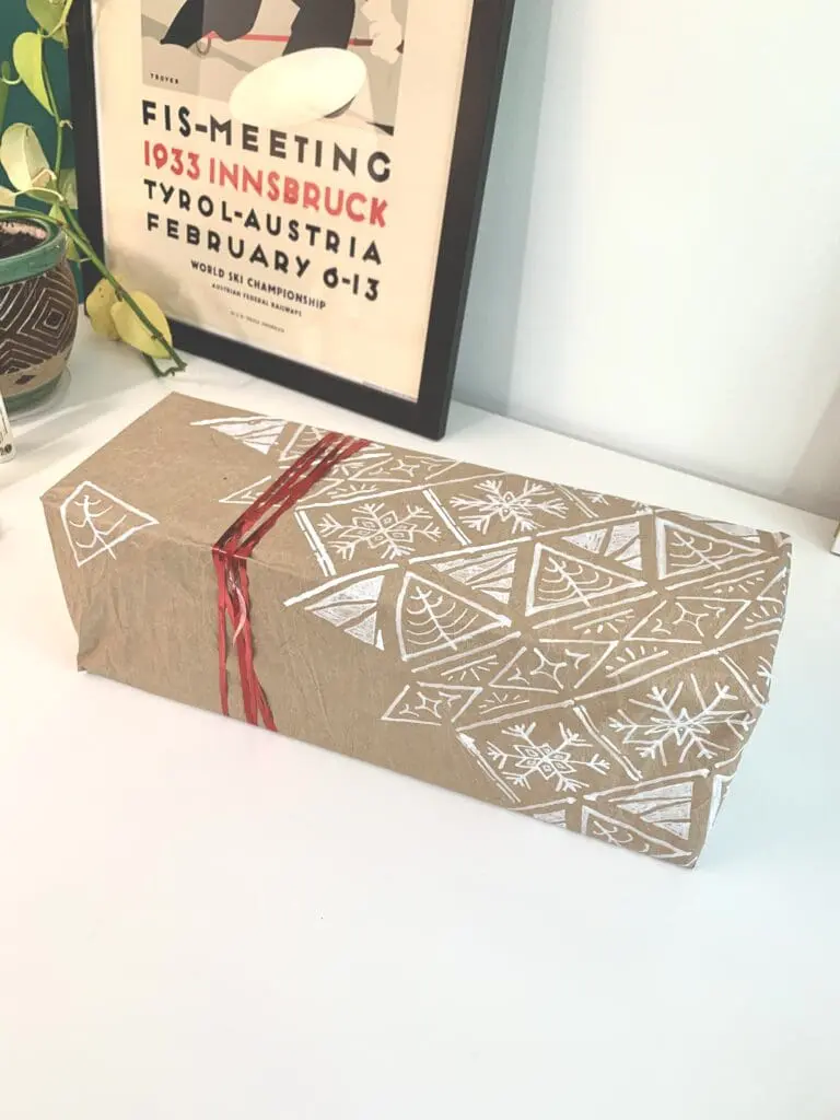 Gift wrapped in brown craft paper with a Christmas pattern stenciled on it in white. The gift has a red ribbon