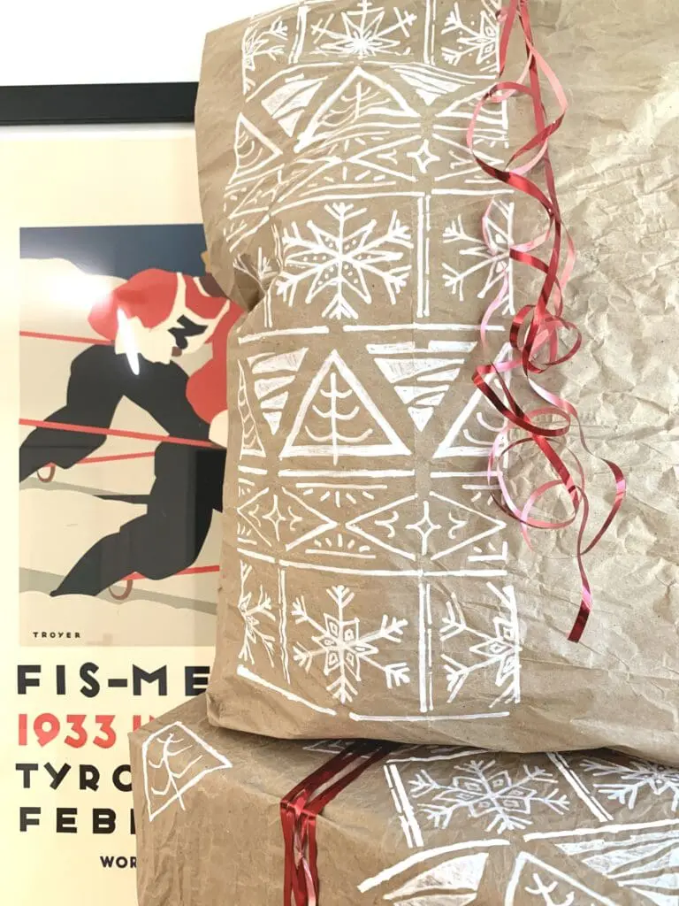 Gift wrapped in brown craft paper with a Christmas pattern stenciled on it in white. The gift has a red ribbon
